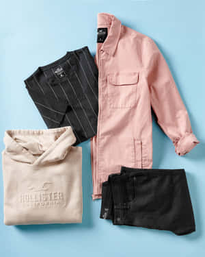 折疊的 Hollister 服裝套裝，包括外套、T 恤、連帽衫和短褲，背景為藍色。 — 海灘度假 壁紙