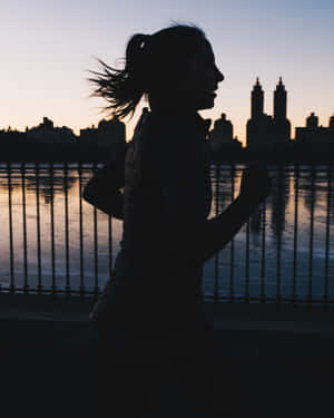 女子在日落時沿著城市海濱跑步，身後是天際線。 — Running 壁紙
