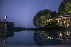 星空倒映在寧靜的無邊際泳池中，旁邊是燈火通明的花園亭閣。 — 牙買加海灘 壁紙