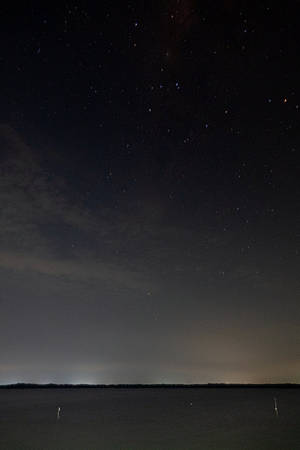 平靜水面上方的星空夜空與遠處的地平線燈光。 — 星星 壁紙