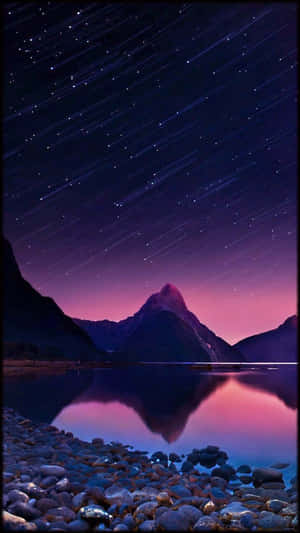 在繁星點點的紫色夜空下，山峰倒映在湖中，湖邊有岩石岸。 — 夜空 壁紙