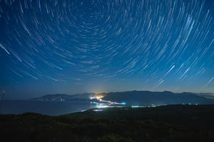 星軌在夜間沿海小鎮上空劃出弧線，遠處燈光沿著海岸線。 — 藍色銀河 壁紙