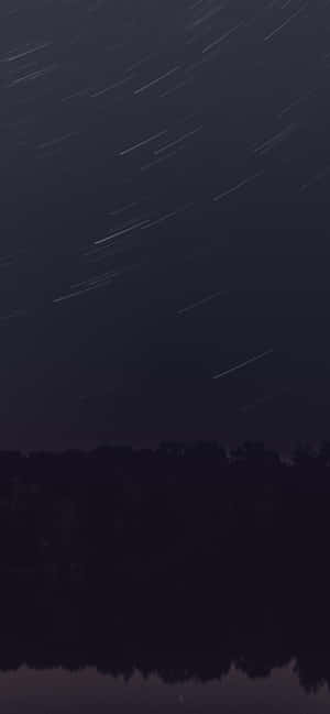 夜晚，星軌在平靜水面上倒映的黑暗森林上方。 — Galaxy A26 壁紙