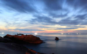 海岸日落，條紋雲彩和寧靜水面上閃爍的遠處燈光。 — 最佳桌面 壁紙
