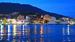 暮色中燈光在水面上閃爍的沿海城市，背景為山脈 — 城市風景 壁紙
