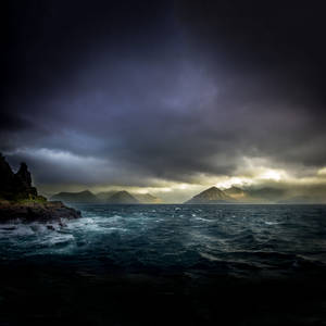 黑暗暴風雨中的海洋，波浪撞擊懸崖，陰沉天空下遠山透過雲層發光。 — 凋零風暴 壁紙