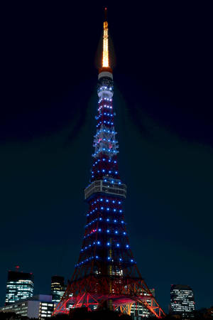 東京塔在夜晚散發紅、藍、金色光芒，被城市建築環繞。 — 日本 壁紙