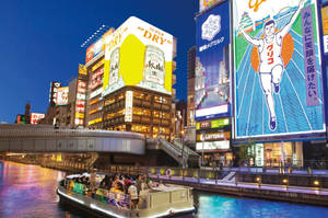 一艘載滿人的運河船在夜間的東京滑過燈火通明的建築物和霓虹招牌。 — 城市 壁紙