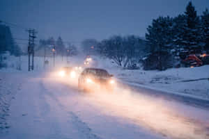 汽車在夜間大雪紛飛的冬季道路上行駛，車頭燈照亮了積雪。 — 冬季風景 壁紙