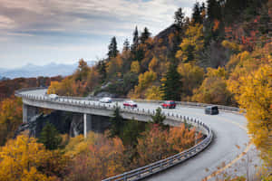 蜿蜒的山路，汽車穿過色彩繽紛的秋樹，駛過一座橋。 — MacBook Air 壁紙