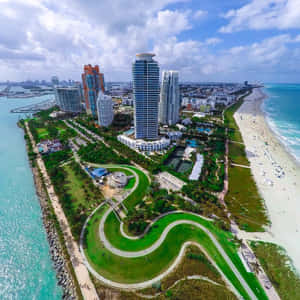 海岸城市，擁有海灘、摩天大樓和蜿蜒的綠色公園，天空部分多雲。 — 邁阿密 壁紙