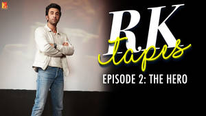 Ranbir Kapoor 身穿白色外套和牛仔褲，交叉雙臂站在「RK Tapes」文字旁。 — 寶萊塢 壁紙