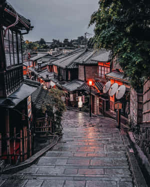 濕滑的石階沿著一條狹窄的日本街道向上延伸，兩旁是傳統木建築，被溫暖的燈籠照亮。 — 日本 壁紙