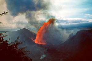 火山爆發，在陰雲密佈的天空下噴出明亮的橙色熔岩和黑煙。 — 自然 壁紙