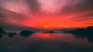 海洋上戲劇性的紅色夕陽，伴隨著黑暗的岩石和平靜的水面 — 海灘日落 壁紙