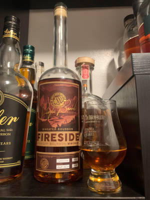 一瓶Fireside Wheated Bourbon Whiskey，旁邊放著品酒杯，置於酒架上。 — 高解析度 壁紙