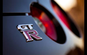 黑色跑車上GT-R標誌的特寫，背景紅色尾燈模糊。 — 日產GTR 壁紙