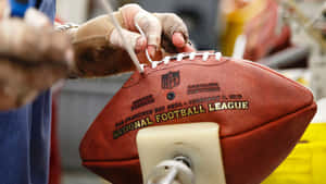 戴手套的手在工場中將鞋帶縫到一個棕色的NFL橄欖球上。 — 足球員 壁紙