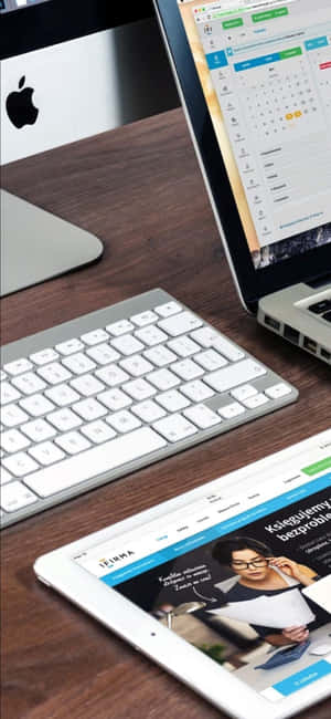 木桌上擺放銀色筆記型電腦、平板電腦和鍵盤，用於數位工作。 — iPhone 18 Pro Max 壁紙