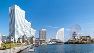 城市天際線，高樓大廈和摩天輪在陽光明媚的日子裡毗鄰河流。 — 橫濱 壁紙