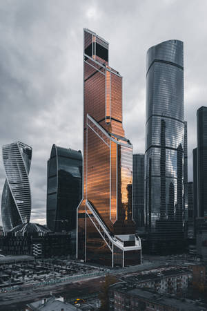 高聳的玻璃摩天大樓在多雲的城市景觀中反射夕陽光線。 — 城市 壁紙