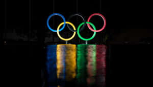 夜晚點亮的奧運五環，在潮濕地面上有彩色倒影。 — 奧運 壁紙
