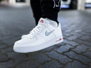 白色Nike Air Force 1運動鞋，紅色和銀色細節，放在鵝卵石人行道上。