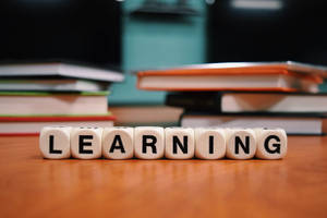 白色字母骰子拼出 learning，放在木桌上，後面有書本。 — 讀書 壁紙