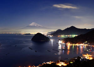 夜晚的富士山，發光的沿海城鎮和星空倒映在平靜的水面上。 — 富士山 壁紙