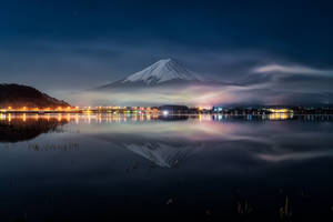 富士山在星空下的平靜湖面倒映，遠處有城市燈光。 — 富士山 壁紙