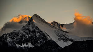 暮色中白雪覆蓋的山峰，金色雲彩在山頂周圍發光。 — MacBook 壁紙