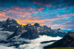 白雪覆蓋的山峰在日落時高聳於雲層之上，天空色彩繽紛 — 美學 Mac 壁紙