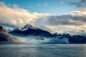 白雪覆蓋的山峰在霧氣瀰漫的湖泊後方升起，天空多雲而戲劇化，金色陽光穿透雲層。 — 藍天 壁紙