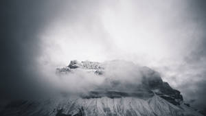 白雪覆蓋的山峰在暴風雨的天空中穿過厚厚雲層聳立。 — 霧濛濛的森林 壁紙