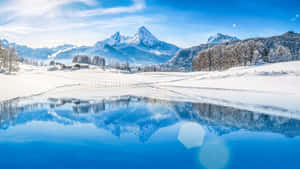 白雪覆蓋的山脈在晴朗的冬日天空下倒映在平靜的藍色湖面上。 — 冬季風景 壁紙