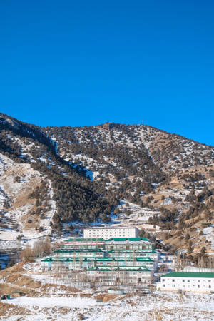 晴朗藍天下，白雪覆蓋的山脈環抱著綠色屋頂的白色建築物。