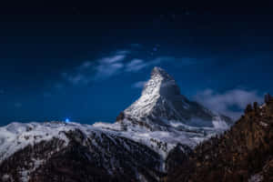 繁星點點的夜空下，白雪覆蓋的馬特洪峰，飄動的雲朵和下方的一盞藍光。 — 夜空 壁紙