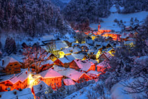 白雪覆蓋的村莊在溫暖的燈光中閃耀，坐落在白雪皚皚的山巒之間，暮色降臨。 — 冬季風景 壁紙