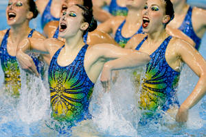 穿著藍色和綠色服裝的韻律泳選手在泳池水中進行充滿活力的動作。 — 運動 壁紙