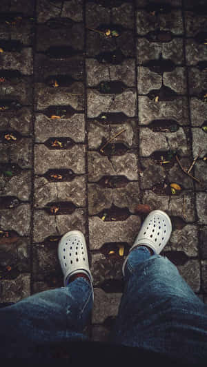 穿著白色Crocs和藍色牛仔褲的人站在石鋪路面上，有落葉。 — Crocs 壁紙