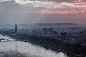 一條河流在黎明時分蜿蜒穿過霧氣瀰漫的城市，背景中有一座高大的紀念碑和山脈。 — 韓國 壁紙