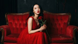 昏暗燈光下，紅裙女性手持紅玫瑰坐在紅色天鵝絨沙發上。 — 珍妮·金 壁紙