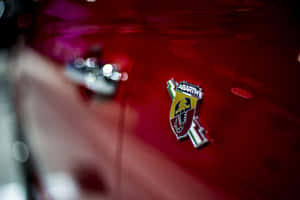 Abarth 汽車徽章在紅色引擎蓋上的特寫，帶有鍍鉻和反射效果。 — Surface Pro 壁紙