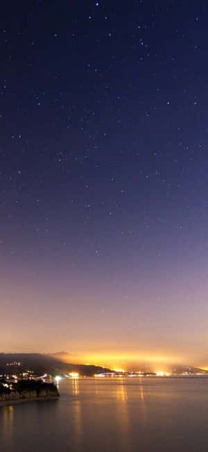 繁星點點的夜空籠罩著平靜的海灣，城市燈光倒映在水面上。 — iPhone Air 壁紙