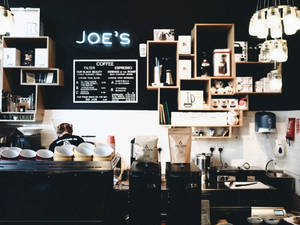 一位咖啡師在現代咖啡店的吧檯後工作，店內有霓虹燈牌和木架。 — 咖啡美學 壁紙