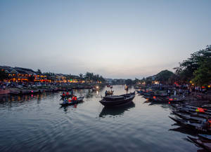 船隻在黃昏時分漂浮在河上，沿岸建築物發出閃爍的燈光。 — 河內 壁紙