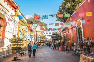 色彩繽紛的 papel picado 橫幅懸掛在熱鬧的行人街上，街上還有歷史建築和購物者。 — 瓜達拉哈拉 壁紙