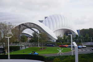 卡丁車在白色未來主義圓頂下的賽道上奔馳，周圍有綠色草坪。 — 音樂錄影帶 壁紙