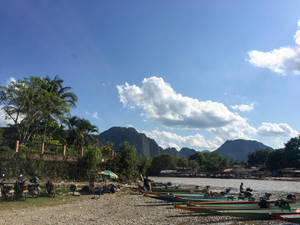 船隻停泊在熱帶河岸，有山脈和棕櫚樹，陽光明媚。 — 曼谷 壁紙