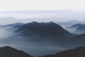 層疊的山峰在蒼白的天空下從薄霧中浮現。 — 乾淨 壁紙